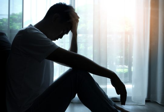 Man silhouetted sitting beside window with head in hands
