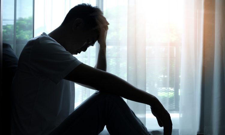 Man silhouetted sitting beside window with head in hands