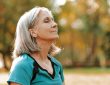 Senior woman wearing sportswear is enjoying a sunny autumn day in the park, closing her eyes and taking a deep breath of fresh air.
