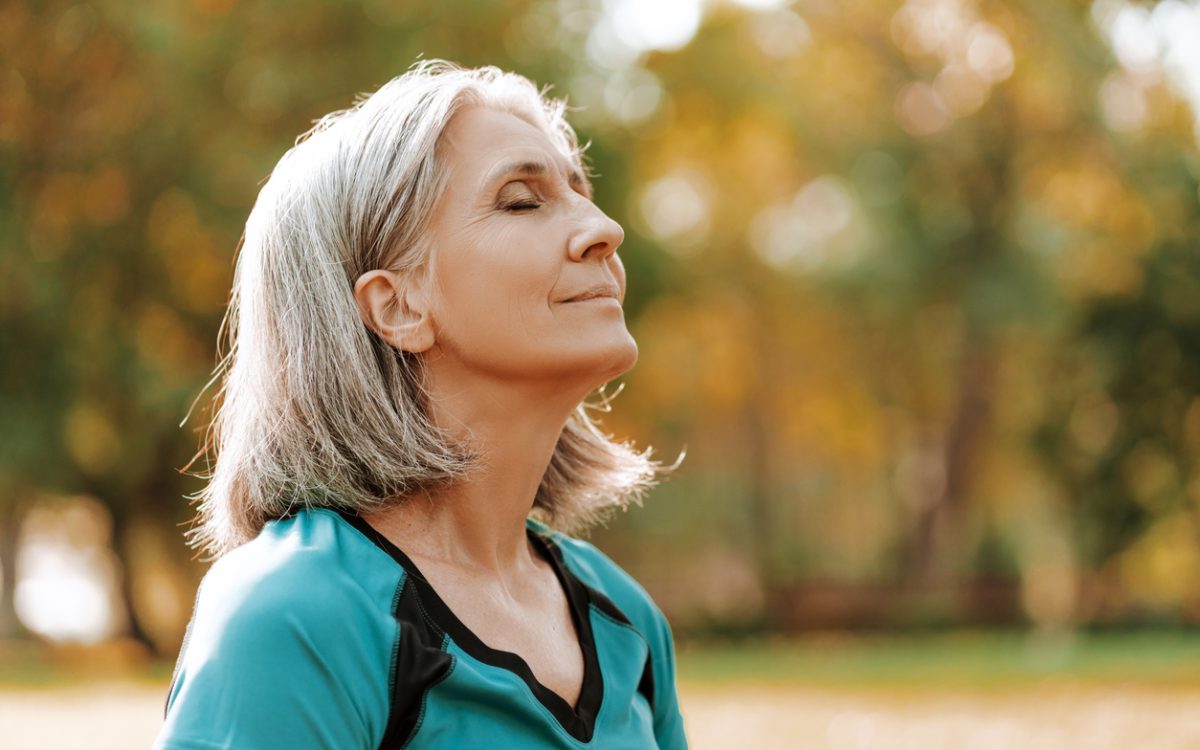Senior woman wearing sportswear is enjoying a sunny autumn day in the park, closing her eyes and taking a deep breath of fresh air.
