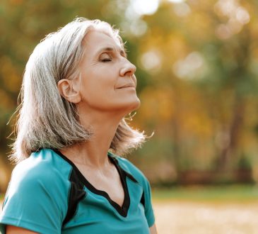 Senior woman wearing sportswear is enjoying a sunny autumn day in the park, closing her eyes and taking a deep breath of fresh air.