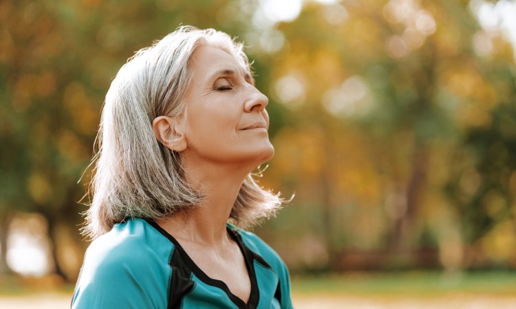 Senior woman wearing sportswear is enjoying a sunny autumn day in the park, closing her eyes and taking a deep breath of fresh air.