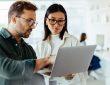 Two people standing and talking while looking at laptop in office