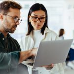 Two people standing and talking while looking at laptop in office