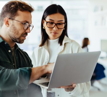 Two people standing and talking while looking at laptop in office