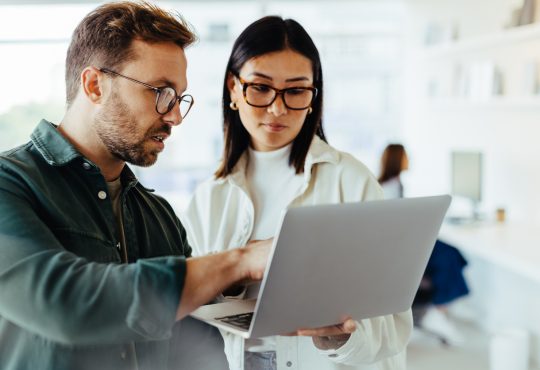 Two people standing and talking while looking at laptop in office