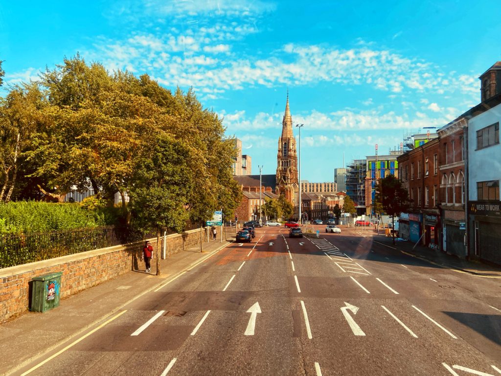 A wide urban street in Belfast with light traffic, flanked by brick buildings and lush green trees.
