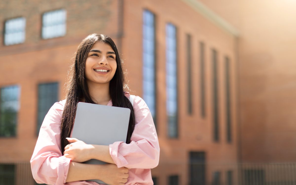Smiling young woman holding laptop outside of school