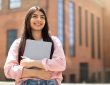 Smiling young woman holding laptop outside of school