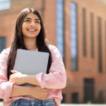 Smiling young woman holding laptop outside of school