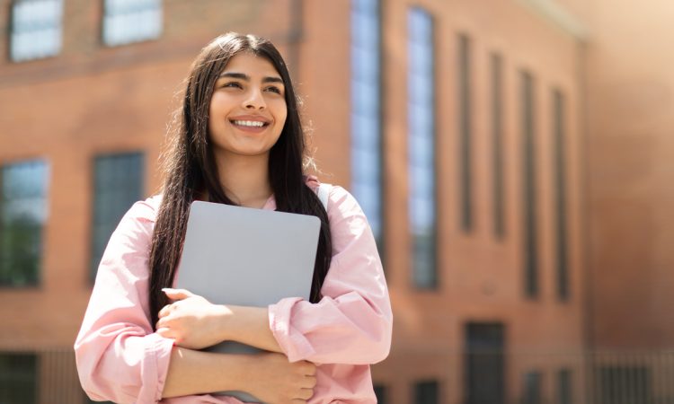 Smiling young woman holding laptop outside of school