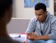 A young african american adult wearing a sweater and jeans, with his elbows on his knees, clasping hands and looking down as he sits on a couch facing a counsellor