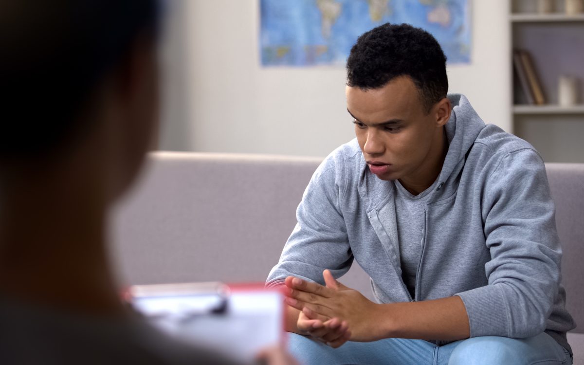 A young african american adult wearing a sweater and jeans, with his elbows on his knees, clasping hands and looking down as he sits on a couch facing a counsellor