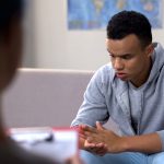 A young african american adult wearing a sweater and jeans, with his elbows on his knees, clasping hands and looking down as he sits on a couch facing a counsellor