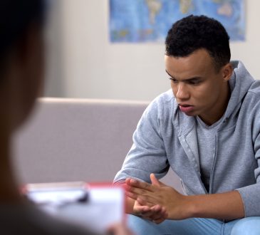 A young african american adult wearing a sweater and jeans, with his elbows on his knees, clasping hands and looking down as he sits on a couch facing a counsellor