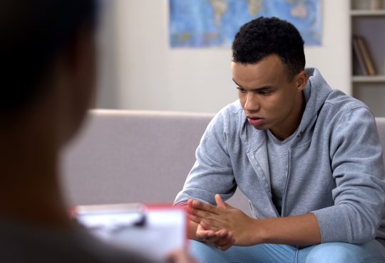 A young african american adult wearing a sweater and jeans, with his elbows on his knees, clasping hands and looking down as he sits on a couch facing a counsellor