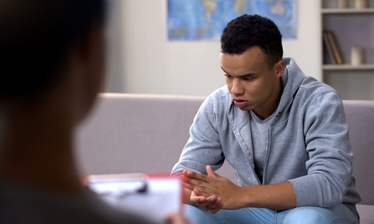 A young african american adult wearing a sweater and jeans, with his elbows on his knees, clasping hands and looking down as he sits on a couch facing a counsellor
