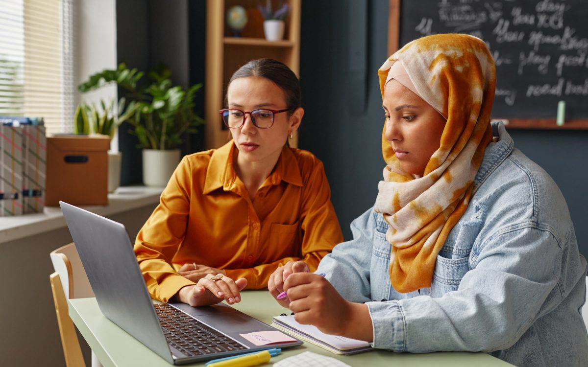 Caucasian young adult woman and Middle Eastern young adult woman wearing hijab learning together in an office, using laptop and taking notes with pens and notebooks