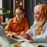 Caucasian young adult woman and Middle Eastern young adult woman wearing hijab learning together in an office, using laptop and taking notes with pens and notebooks