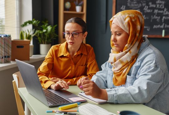 Caucasian young adult woman and Middle Eastern young adult woman wearing hijab learning together in an office, using laptop and taking notes with pens and notebooks
