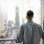Rear view of young man standing on balcony, gazing out at modern urban landscape filled with high-rise buildings and busy roads, taking moment to admire big city view from his new, luxury apartment