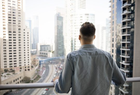 Rear view of young man standing on balcony, gazing out at modern urban landscape filled with high-rise buildings and busy roads, taking moment to admire big city view from his new, luxury apartment