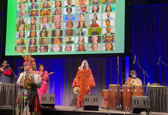 An image of the cultural celebration at Cannexus26 with indigenous music performers holding instruments on a stage. Behind them is a blue curtain and a slide showing photos of CERIC board and staff headshots.
