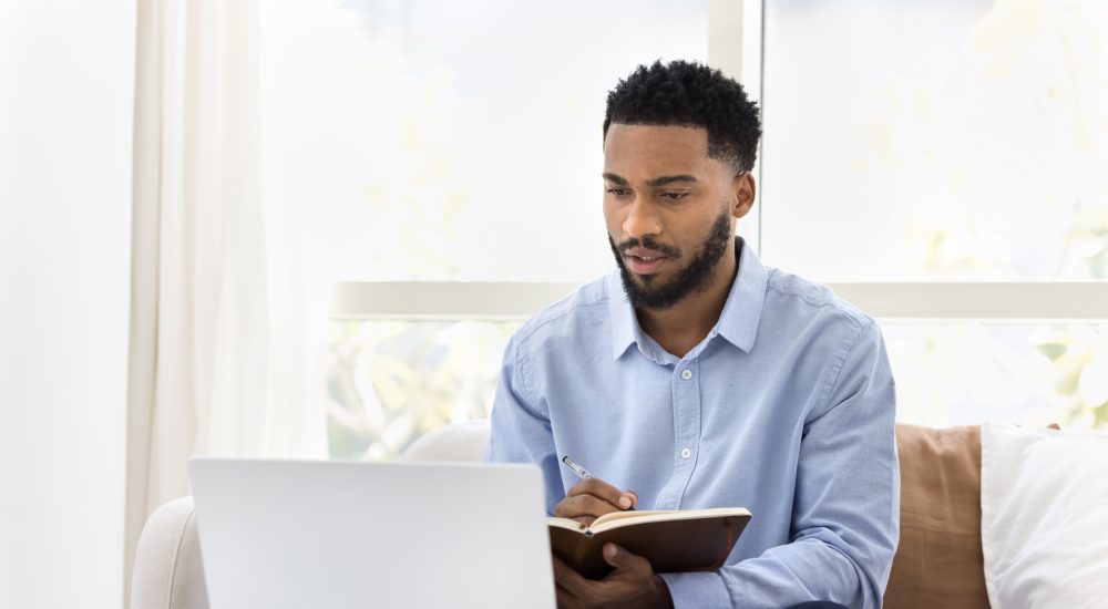 Serious African student man watching online lecture, training seminar on laptop at home, writing notes, summary of webinar, studying on Internet, doing learning homework, speaking on video call