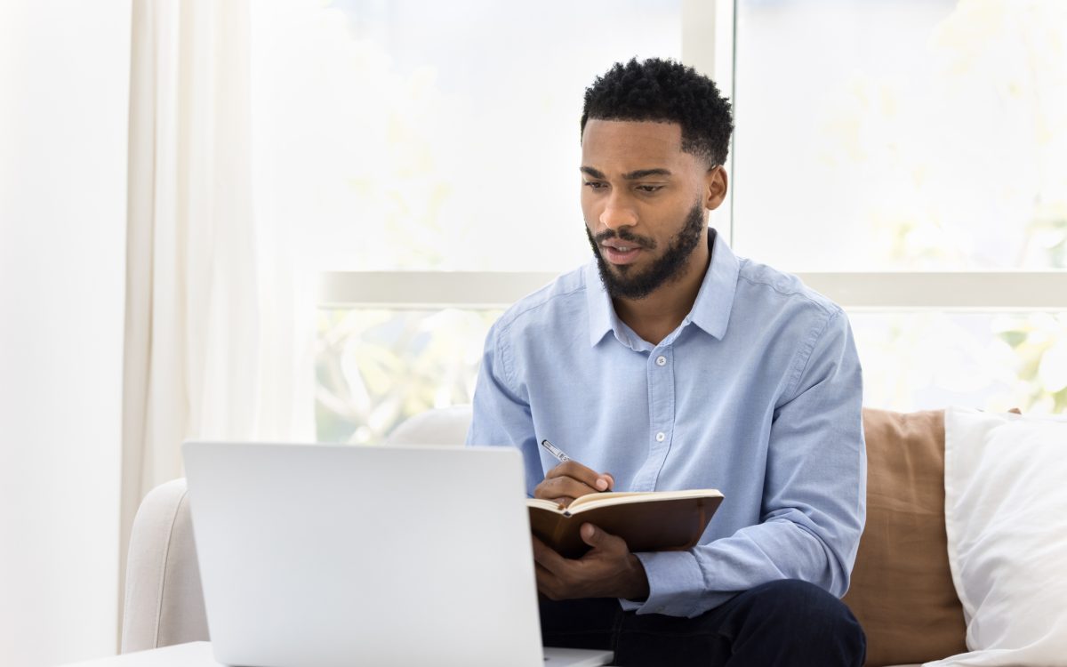 Serious African student man watching online lecture, training seminar on laptop at home, writing notes, summary of webinar, studying on Internet, doing learning homework, speaking on video call