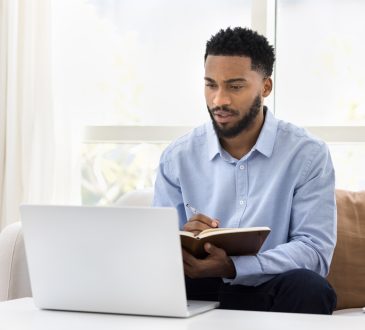 Serious African student man watching online lecture, training seminar on laptop at home, writing notes, summary of webinar, studying on Internet, doing learning homework, speaking on video call