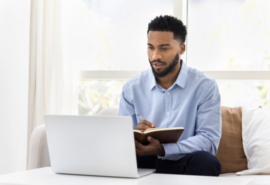 Serious African student man watching online lecture, training seminar on laptop at home, writing notes, summary of webinar, studying on Internet, doing learning homework, speaking on video call