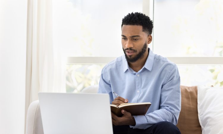 Serious African student man watching online lecture, training seminar on laptop at home, writing notes, summary of webinar, studying on Internet, doing learning homework, speaking on video call