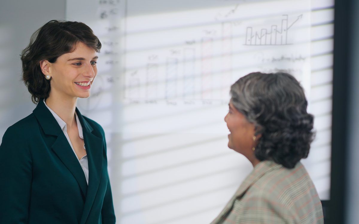 European Caucasian young adult gen z diverse business woman lady standing talk workday task indoor job office place. Teen female worker boss manager make conversation give advice helping consulting discuss client insurance benefit