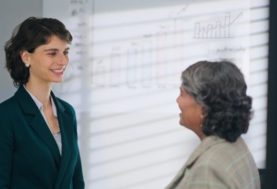 European Caucasian young adult gen z diverse business woman lady standing talk workday task indoor job office place. Teen female worker boss manager make conversation give advice helping consulting discuss client insurance benefit