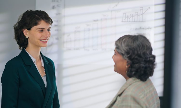 European Caucasian young adult gen z diverse business woman lady standing talk workday task indoor job office place. Teen female worker boss manager make conversation give advice helping consulting discuss client insurance benefit