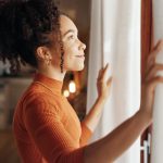 Curtains, happy woman and thinking of future in home, planning and reflection of mindset with view. Window, smile and African girl with vision, ideas and dream for morning inspiration in living room