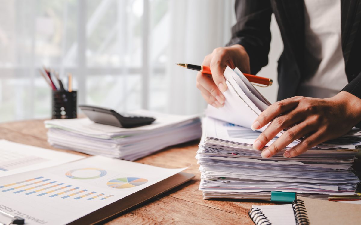 A desk with piles of reports being handled by a set of hands holding a pen. The desk is by a well-lit window, and one pile of reports has a calculator on it.