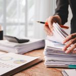 A desk with piles of reports being handled by a set of hands holding a pen. The desk is by a well-lit window, and one pile of reports has a calculator on it.