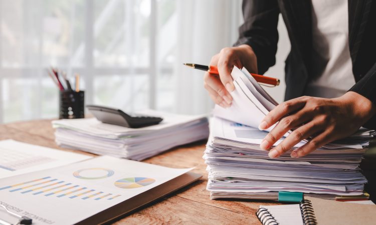 A desk with piles of reports being handled by a set of hands holding a pen. The desk is by a well-lit window, and one pile of reports has a calculator on it.