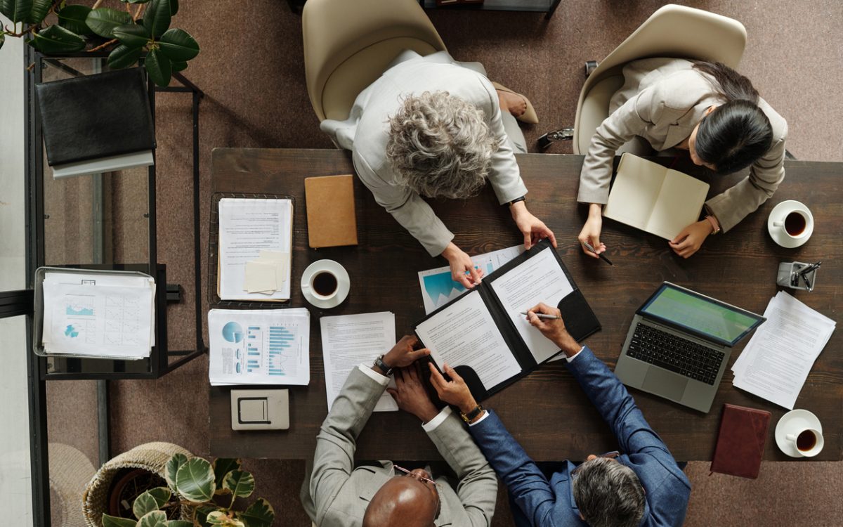 Group of middle aged multiethnic business professionals collaborating around table, reviewing documents and using laptop, top view showing teamwork and corporate meeting environment