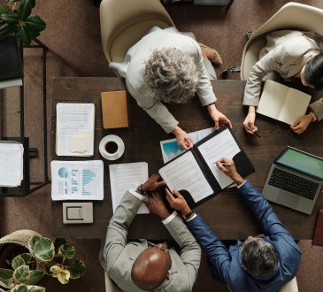 Group of middle aged multiethnic business professionals collaborating around table, reviewing documents and using laptop, top view showing teamwork and corporate meeting environment