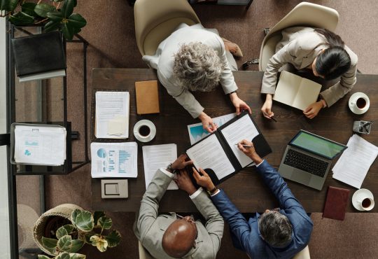 Group of middle aged multiethnic business professionals collaborating around table, reviewing documents and using laptop, top view showing teamwork and corporate meeting environment