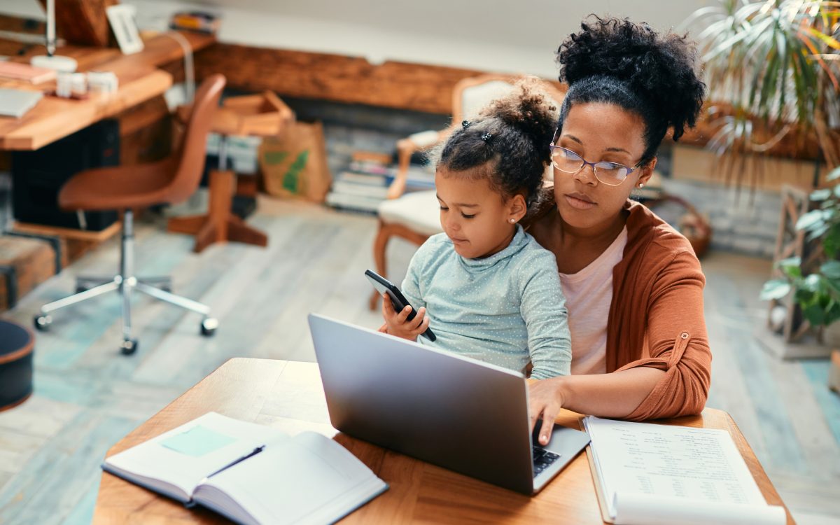 African American stay at home mother working on laptop while daughter is sitting on her lap and using smart phone.