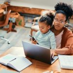 African American stay at home mother working on laptop while daughter is sitting on her lap and using smart phone.