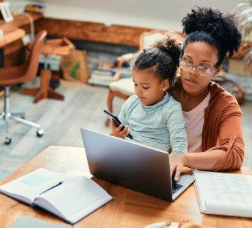 African American stay at home mother working on laptop while daughter is sitting on her lap and using smart phone.