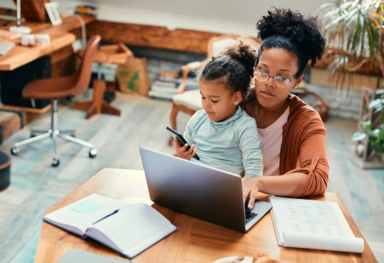 African American stay at home mother working on laptop while daughter is sitting on her lap and using smart phone.