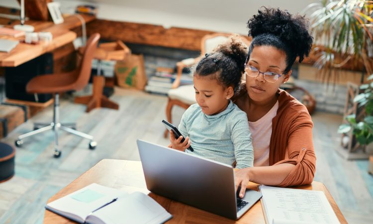 African American stay at home mother working on laptop while daughter is sitting on her lap and using smart phone.