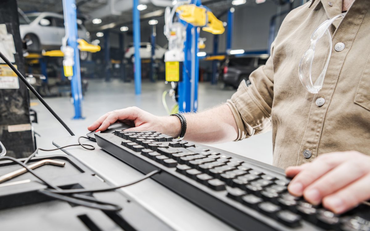 Caucasian Automotive Technician Checking Vehicle Database Documentation. Hands on Computer Keyboard Inside Auto Service Station.