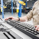 Caucasian Automotive Technician Checking Vehicle Database Documentation. Hands on Computer Keyboard Inside Auto Service Station.
