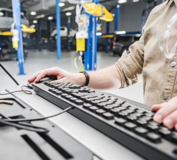 Caucasian Automotive Technician Checking Vehicle Database Documentation. Hands on Computer Keyboard Inside Auto Service Station.